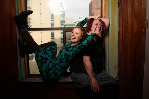 Two people sit perched in a windowsill.  Ingrid, short red hair, wears and green and black dress and is leaning back against Kathleen, playfully poking at Kathleen's face and smiling.  Kathleen, curly purple hair, wears a black shirt and leans against the window with her elbow.  Photo by Helene Cyr.